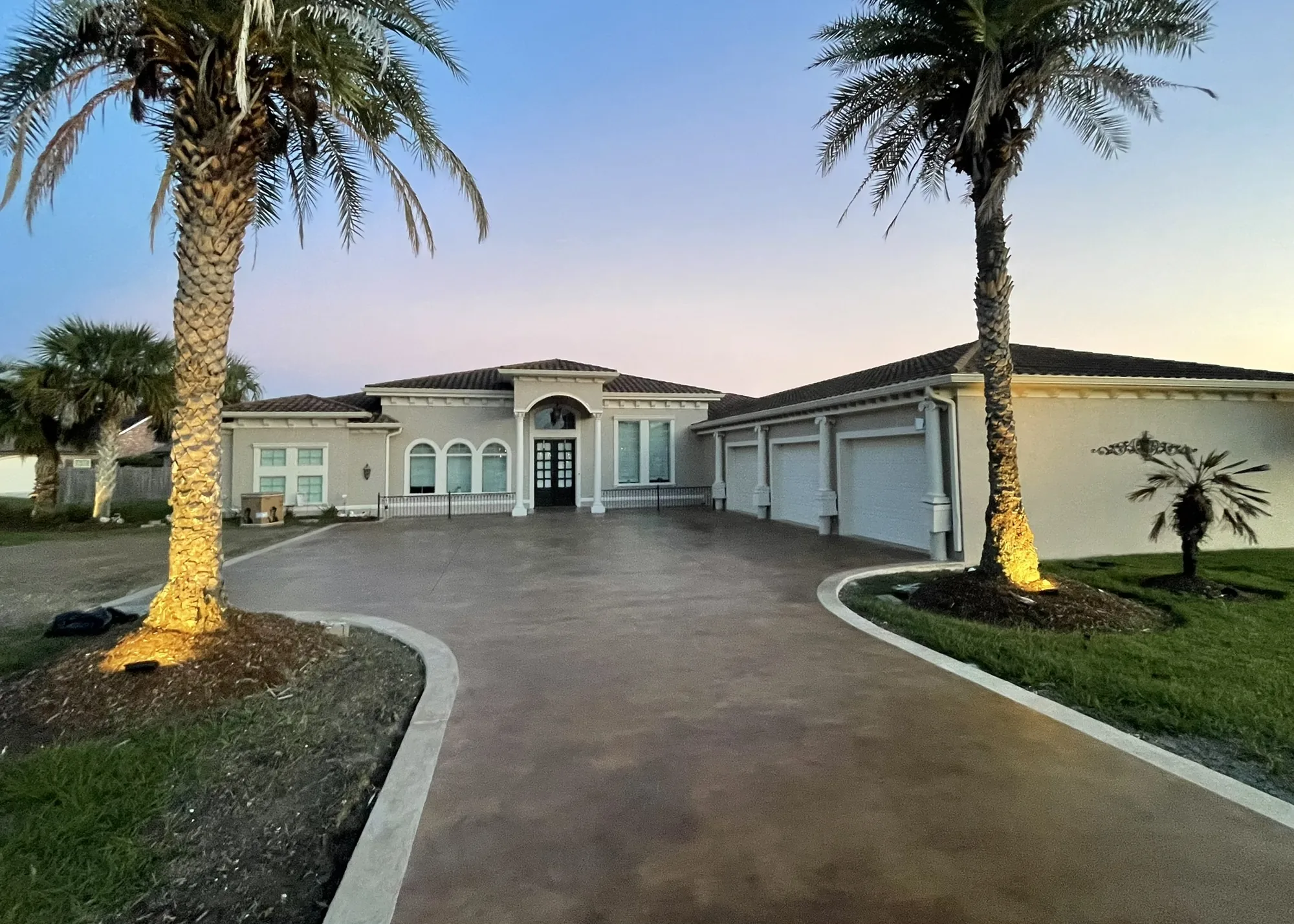 Curved decorative concrete driveway leading to a stucco home at dusk, framed by lit palm trees.