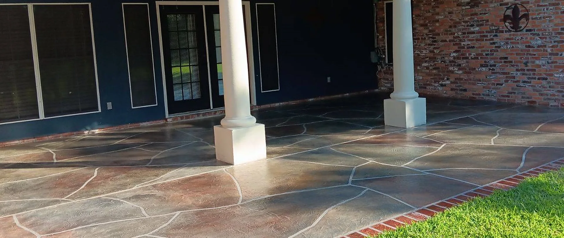 Covered patio finished in a multi-tone flagstone pattern with a brick border, between white columns and a brick wall.